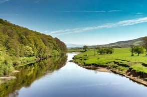 A view of the River Lune near Lancaster on a sunny day, with green fields and wooded slopes.